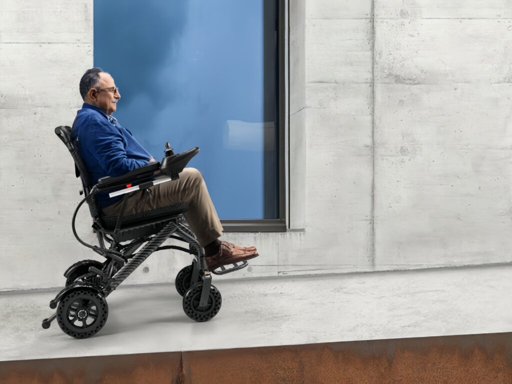A senior man riding a lightweight, foldable electric wheelchair with a joystick control, climbing an outdoor ramp next to a concrete wall.